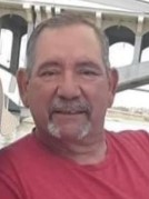 Photo of a middle-aged man with tanned skin and short gray hair and beard. He is wearing a red shirt. 