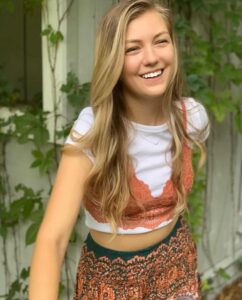 Photo of a young woman with fair skin and long blond hair, wearing an orange patterned skirt and top that barely shows her midriff. She is smiling for the camera.