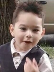 Photo of a young boy with light skin and brown hair which is sticking up from his head. He is wearing a white collared shirt and gray vest. He has his mouth open in amusement.