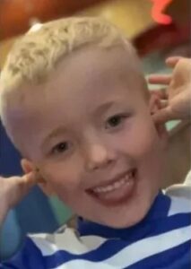 Photo: A boy with short, curly blond hair and fair skin, smiling, holding his thumbs to his ears and wiggling his fingers; he is wearing a blue and white striped shirt.