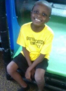 Photo: Young boy, seated, with deep brown skin, wide nose, and broad smile, his black hair cut short. He is wearing black shorts and a yellow T-shirt printed with the words Gotta Catch 'Em All.