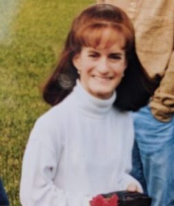 Photo: Mary Mount is a short woman with auburn hair cut in bangs. Her skin is pale; she is wearing a white turtleneck. The photo is taken outdoors. She is smiling.