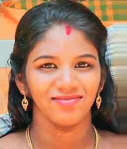 Photo of a young woman with tan skin, her dark-brown hair pulled back from her face. She is wearing a golden necklace and golden earrings, and there is a bindi of red paint on her forehead. She is smiling for the camera.