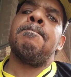 Photo: An African-American man, close-up, taken from below. He has a scruffy beard and mustache. One of his eyes points off to the left. He is wearing a sports jersey with a yellow collar.