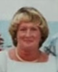 Blurry photo of a middle-aged woman with graying sandy hair, round face,  and pale skin; she wears a white top and pearl necklace.