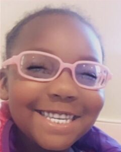 Photo: Young girl with brown skin and pink glasses, smiling for the camera, eyes squeezed shut in laughter.