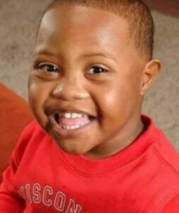 Photo: A young boy with dark skin and very short black hair, wearing a red shirt, smiling for the camera.