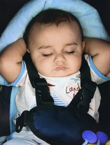 Photo: A baby with dark hair and olive-toned skin, wearing a whtie-and-blue onesie, belted into a blue car seat with a five-point belt. His hands are propped behind his head, and his eyes closed; he looks relaxed.