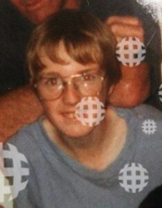 Photo: A thin teenage boy with light skin and curly light-brown hair, wearing large glasses and a baggy gray T-shirt.