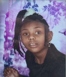 Photo of an African-American girl with medium-brown skin and brown eyes, wearing a black top. Her hair is done in small, neat braids and gathered into a scrunchie at the top of her head.