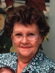 Photo: A middle-aged woman with fair skin and short, curly brown hair. She is wearing glasses and a blue flowered blouse.