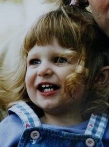 Photo of a toddler girl in blue overalls and white blouse, her dark-blond hair cut in bangs. She has fair skin and is smiling, showing her baby teeth.
