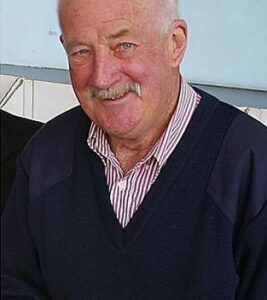An older man with ruddy skin and white hair and mustache, wearing a cardigan and button-up shirt.