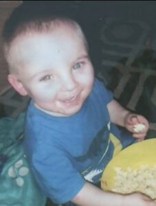 Photo: A toddler boy sits with a bowl of popcorn in his lap, holding a piece in one hand. He has fair skin and brown hair, and is wearing a blue T-shirt. He is smiling for the camera.