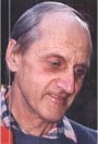 Photo of an elderly man, balding with gray hair and light skin. He has a thin face, and is looking down. He is wearing a checked shirt.