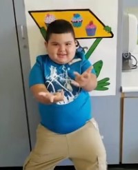  Photo of a small, chubby boy with short brown hair and fair skin; he is wearing a blue T-shirt and khakis. He has his hands out in front of him, waving them or perhaps dancing. 