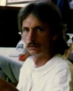 Photo: A man with light skin and dark-brown hair and mustache, wearing a white shirt, expression neutral. His hair is shoulder-length.