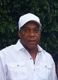 Photo: A dark-skinned man wearing a white baseball cap and white button-down shirt; his expression is neutral, and there is a tree in the background.