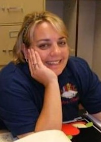 Photo: A woman with fair skin and blond hair pulled back in a ponytail, sitting at a desk, smiling for the camera. She is wearing a blue T-shirt, and leaning her head in one hand.