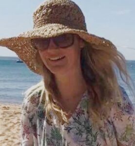 Photo: A smiling woman standing on a beach, wearing sunglasses, a straw hat, and a colorful shirt. She has fair skin and long blonde hair.