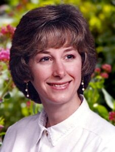 Photo of a woman with fair skin and short, wavy brown hair styled to frame her face; she is smiling for the camera. She is wearing white pearl drop earrings and a neatly buttoned white blouse.