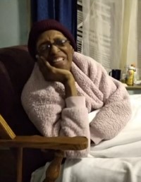 Photo: An older woman with brown skin and glasses, reclining in a wooden chair with red upholstery. She is wearing a fluffy pink robe and a maroon beanie hat, and is covered with a white blanket. She is smiling for the camera, head propped on one hand.