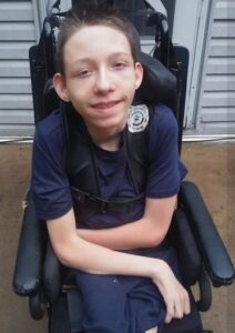 A fair-skinned, brown-haired boy sits in a wheelchair; he is wearing navy blue shirt and pants, and is smiling for the camera. 