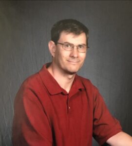 Portrait photo of a man with fair skin, brown hair, glasses, and a red shirt, smiling for the camera.