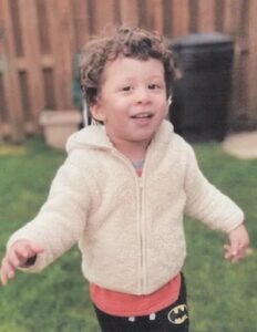 Photo of a young boy with brown curly hair, brown eyes, and pale-brown skin, wearing a fleecy sweatshirt and pants with a Batman logo. The photo is taken outdoors, and he is smiling and waving his hands.