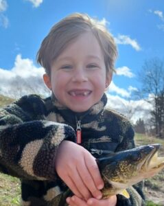 Photo of a boy with fair skin and light-brown hair, wearing a hoodie. He is holding a fish and smiling, showing his missing front teeth.