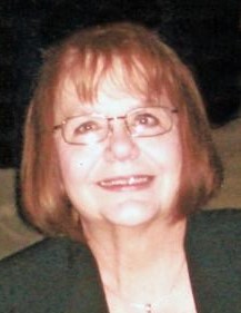 Photo of an older woman with pale skin and red, chin-length hair, wearing glasses and a dark-green blazer.