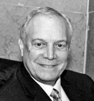 Black and white photo of a man in late middle age, gray-haired with a receding hairline. He is wearing a suit. He has light skin, and is smiling. 