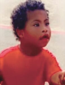 Photo of a toddler boy with curly black hair and light-brown skin, his mouth slightly open; he is wearing an orange T-shirt.