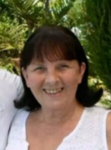 Photo of a fair-skinned woman with dark-brown hair cut in bangs and curled at the tip. She is smiling for the camera. She is wearing a white tank top.