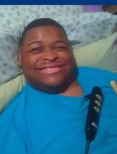 Photo of a young African-American manlying propped up in bed, smiling. He has a hospital call button lying across his chest. He is wearing a teal shirt.