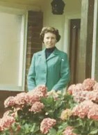 Old-fashioned photo of a woman with brown curly hair and fair skin, wearing a blue jacket, smiling for the camera. There are pink flowers in the foreground.