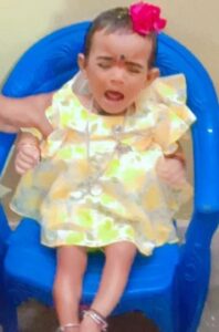 Photo of a baby sitting in a blue chair, wearing a yellow dress and pink flower in her hair. She has light-brown skin, short dark-brown hair, and a bindi on her forehead. She seems upset, her eyes shut and her mouth open in a cry. Off to the side, an adult is holding her arm. 