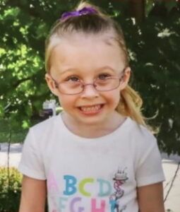Photo of a young girl, fair-skinned and blond, her hair in a ponytail, wearing glasses and a white T-shirt printed with a colorful alphabet.