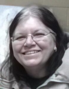 Photo of a middle-aged woman with brown hair growing out into gray roots, wearing glasses; she has pale skin, and is smiling for the camera.