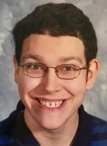 Portrait photo of a young man smiling for the camera; he has curly brown hair and is wearing glasses.