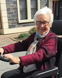 Photo of a white-haired older woman with pink, fair skin and glasses, wearing a burgundy sweater and scarf, hands on the controls of a mobility scooter.