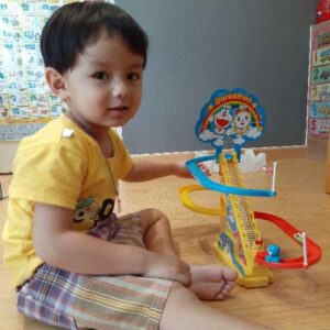 Photo of a small boy with light-brown skin and short black hair, wearing a yellow T-shirt and striped gray and white shorts, sitting on the floor next to a colorful toddler toy.