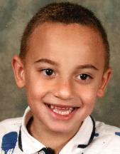 Photo of a smiling boy with light-brown skin and dark-brown eyes, wearing a white striped polo shirt. His brown hair is cut short.