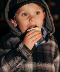 Photo of a light-skinned boy with blue eyes, wearing a gray hoodie and eating a cupcake with white frosting.