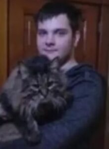 Photo of a young man with short brown hair and fair skin, wearing a gray sweater, holding a large, fluffy tabby cat.