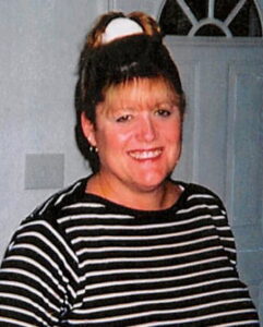 Photo of a middle-aged woman with dark-blond hair and fair skin, smiling for the camera. Her hair is pulled back in a ponytail and cut into bangs; she is wearing a black and white striped sweater.