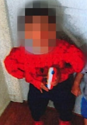 Photo of a boy with brown skin and black hair, wearing a red sweater and black pants. His face is blurred out. 