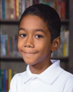 Photo of a boy with deep brown eyes, light-brown skin, and short brown hair, wearing a white polo shirt.