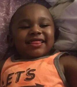 Photo of a young boy with dark skin, grinning at the camera, lying back on a pillow, wearing an orange tank top printed with "Let's" (the rest of the message is out of frame).