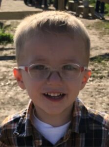 Photo of a small fair-skinned boy with short blond hair, smiling for the camera, wearing plastic-rimmed glasses and a flannel shirt over a white T-shirt.
